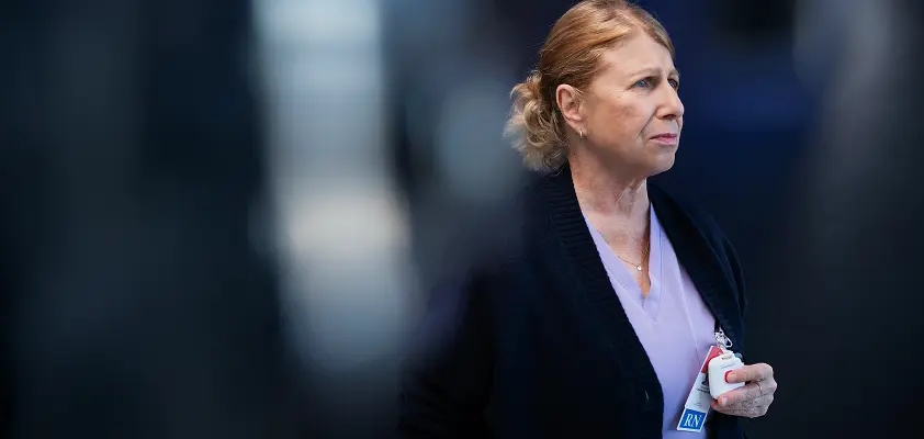 Middle-aged nurse in lavender scrubs and a dark cardigan stands in a hospital corridor, holding her Kontakt io badge and looking pensively to the side, with a softly blurred clinical background.